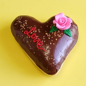 A beautifully decorated Valentine’s Love Heart Donut, filled with strawberry jam, topped with a smooth chocolate glaze, adorned with gold sprinkles, edible star accents, and a delicate candy rose, featuring a hand-piped ‘Love’ design. A perfect romantic treat for Valentine’s Day.