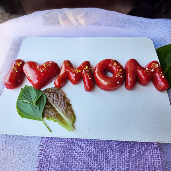 Strawberry mirror glaze donuts spelling “I ❤️ MOM,” decorated with edible gold powder, sugar pearls, and gold sprinkles for Mother’s Day.